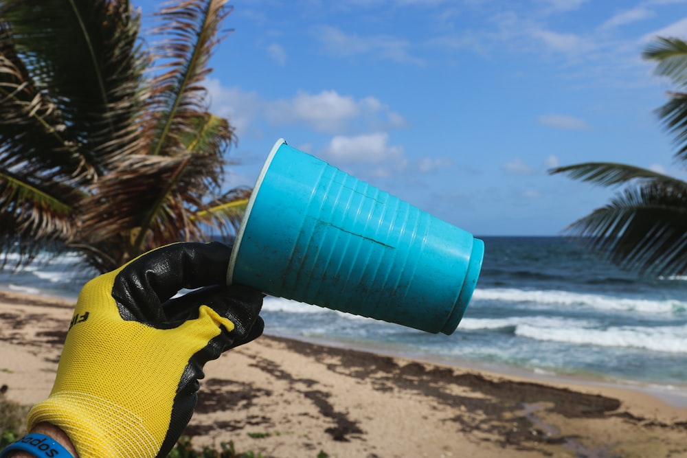 blauer Plastikbecher, der mit gelben Handschuhen am Strand in der Hand gehalten wird