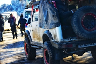 An off-road vehicle with large tires and a spare wheel mounted at the back is parked on a muddy ground. The vehicle appears worn, with visible dirt and wear marks. In the background, a few people are standing and interacting near other vehicles. The scene is set in a cold, possibly snow-covered environment, indicated by the attire of the people and the distant snowy landscape.