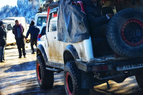 An off-road vehicle with large tires and a spare wheel mounted at the back is parked on a muddy ground. The vehicle appears worn, with visible dirt and wear marks. In the background, a few people are standing and interacting near other vehicles. The scene is set in a cold, possibly snow-covered environment, indicated by the attire of the people and the distant snowy landscape.