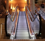 A modern escalator with sleek metal railings and glass sides, illuminated by warm overhead lights. The setting includes adjacent stairs and a polished wood ceiling, enhancing the contemporary architecture.