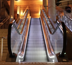 A modern escalator with sleek metal railings and glass sides, illuminated by warm overhead lights. The setting includes adjacent stairs and a polished wood ceiling, enhancing the contemporary architecture.