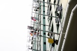 A tall building with multiple rows of clothes hanging on poles protruding from the windows. The clothing is colorful and varied, suggesting it is laundry hung out to dry. The building's facade is light-colored with rectangular windows, some of which are open.