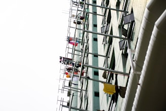 A tall building with multiple rows of clothes hanging on poles protruding from the windows. The clothing is colorful and varied, suggesting it is laundry hung out to dry. The building's facade is light-colored with rectangular windows, some of which are open.