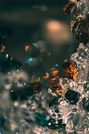 A close-up view of multifaceted gemstones, possibly crystals, reflecting light with prismatic color patterns. The stones are clear with hints of amber and teal tones, creating a sparkling effect against a dark blurred background.