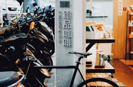A street view captures a row of parked scooters on one side and a cozy cafe interior on the other. The cafe features wooden decor, with a rack holding coffee-related items and a bicycle parked prominently outside. There is a vertical sign displaying characters, likely indicating the name or type of establishment.