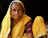 An elderly woman wearing a vibrant yellow sari with green and red patterned accents is seated. She has deep wrinkles on her face, and her expression is contemplative. Her clothing is traditional and richly colored, standing out against a dark background.