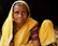 An elderly woman wearing a vibrant yellow sari with green and red patterned accents is seated. She has deep wrinkles on her face, and her expression is contemplative. Her clothing is traditional and richly colored, standing out against a dark background.