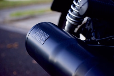 A close-up view of a motorcycle's black exhaust pipe with the logo 'Vance & Hines Competition' clearly visible. The image captures the shiny metallic parts and a coil spring from the motorcycle's suspension system in the background.
