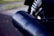 A close-up view of a motorcycle's black exhaust pipe with the logo 'Vance & Hines Competition' clearly visible. The image captures the shiny metallic parts and a coil spring from the motorcycle's suspension system in the background.