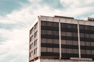 A modern office building with a grid of large, dark-tinted windows divided by concrete panels. The structure appears to be mid-rise with a clean, minimalist design. Surrounding the building is a sky with wispy clouds, giving the scene a serene atmosphere.