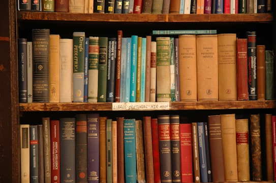 A wooden bookshelf filled with an assortment of books of various sizes and colors. The spines of the books are visible, displaying an array of titles and authors. Most books appear to have hard covers, and there is a mix of older and newer volumes. The bookshelf has three visible shelves, and a label on the middle shelf reads 'LIBROS EDITORIAL MIR MOSC&Uacute;'.