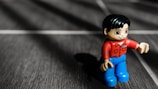 A small toy figure is positioned on a textured surface, casting a shadow. The toy has a red shirt, blue pants, and a smiling expression. The surface has a dark, wooden appearance with white lines creating a grid pattern.