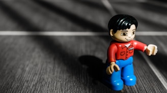 A small toy figure is positioned on a textured surface, casting a shadow. The toy has a red shirt, blue pants, and a smiling expression. The surface has a dark, wooden appearance with white lines creating a grid pattern.