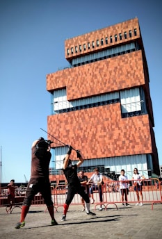 Two individuals are engaged in a mock sword fight in front of a modern, red-bricked building. The participants wear protective gear, including padded gloves. A small crowd watches the duel, standing behind a red barricade. The building behind features large glass windows and a unique, stacked design.