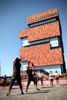 Two individuals are engaged in a mock sword fight in front of a modern, red-bricked building. The participants wear protective gear, including padded gloves. A small crowd watches the duel, standing behind a red barricade. The building behind features large glass windows and a unique, stacked design.