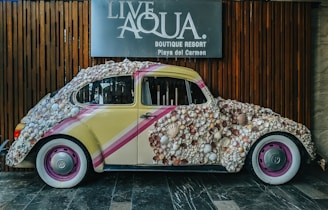 A vintage Volkswagen Beetle car is covered in seashells, creating an artistic installation. The car has a yellow base with diagonal pink and white stripes, and the hubcaps are painted pink. It is parked against a wooden background, and a sign for 'Live Aqua Boutique Resort Playa del Carmen' is visible above the car.