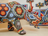 A colorful beaded rhinoceros sculpture with intricate geometric patterns and vibrant designs, displayed alongside similar artistic works on the wall. The beads form detailed images and motifs in a variety of colors, including reds, blues, yellows, and greens.