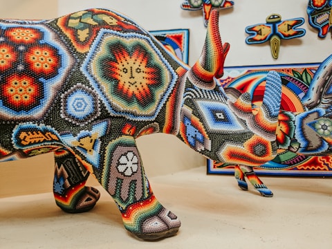 A colorful beaded rhinoceros sculpture with intricate geometric patterns and vibrant designs, displayed alongside similar artistic works on the wall. The beads form detailed images and motifs in a variety of colors, including reds, blues, yellows, and greens.