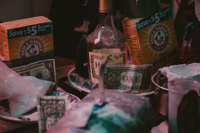 A close-up of a cluttered kitchen or tabletop scene featuring boxes of Arm & Hammer baking soda, a bottle labeled 'Hennessy,' and multiple U.S. one-dollar bills. A few plates, some with white powder, and various containers are also visible.