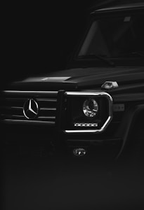 A close-up view of the front of a luxury SUV focusing on the headlight and part of the grille. The image is in black and white, highlighting the sleek and modern design. The SUV features a prominent logo and clean lines, set against a dark background, emphasizing the vehicle's bold and robust appearance.