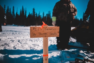 A wooden sign with handwritten text referencing a price increase by tariffs is planted in the snow. People are gathered around in winter clothing, and a snowmobile is visible. The background features a forest of evergreen trees under a clear blue sky.