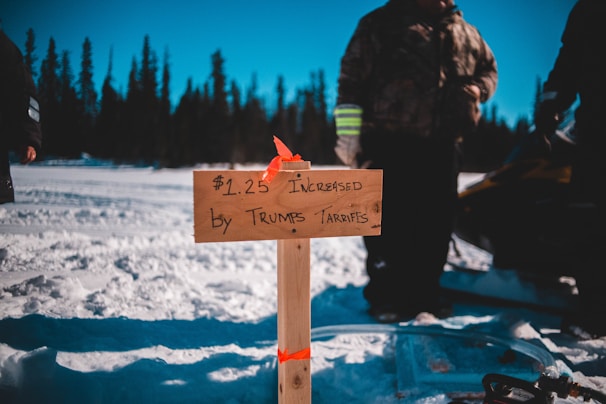 A wooden sign with handwritten text referencing a price increase by tariffs is planted in the snow. People are gathered around in winter clothing, and a snowmobile is visible. The background features a forest of evergreen trees under a clear blue sky.