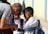 A group of children in school uniforms gather around an adult who is showing them something in his hands. The scene appears outdoors, with a sunny, clear day evident from the shadows and lighting. The children have backpacks, suggesting they are on their way to or from school.