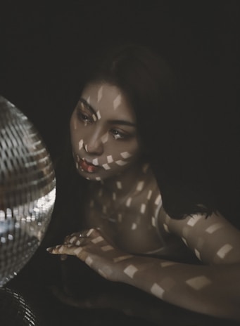 A person gazes thoughtfully at a reflective disco ball, with patterned light casting geometric shadows across their face and body. The overall setting is dark, emphasizing the play of light and shadow.