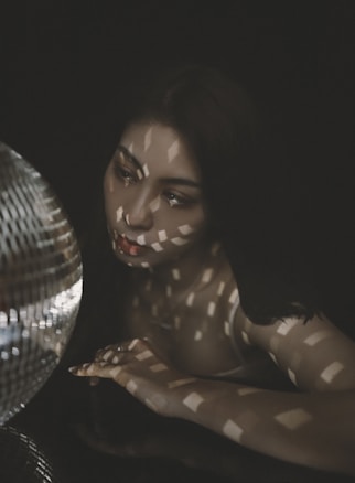 A person gazes thoughtfully at a reflective disco ball, with patterned light casting geometric shadows across their face and body. The overall setting is dark, emphasizing the play of light and shadow.