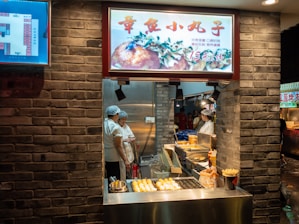 A street food stall showcases a variety of snacks, with two workers wearing hair nets engaged in conversation. The stall has a brick façade, and there are cooking appliances such as fryers visible. An illuminated menu features an image of food above the cooking area. Bright lighting enhances the visibility of the food displayed.