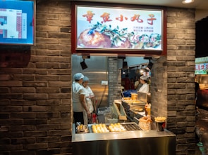 A street food stall showcases a variety of snacks, with two workers wearing hair nets engaged in conversation. The stall has a brick façade, and there are cooking appliances such as fryers visible. An illuminated menu features an image of food above the cooking area. Bright lighting enhances the visibility of the food displayed.