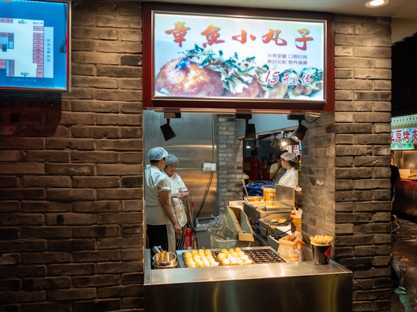 A street food stall showcases a variety of snacks, with two workers wearing hair nets engaged in conversation. The stall has a brick façade, and there are cooking appliances such as fryers visible. An illuminated menu features an image of food above the cooking area. Bright lighting enhances the visibility of the food displayed.