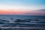 A serene ocean scene at dusk with gentle waves and a horizon tinted with shades of orange, pink, and blue. The sky is vast and lightly clouded, creating a tranquil atmosphere over the calm water.