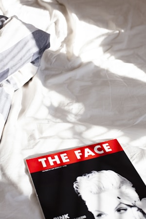 A magazine with a red and white cover lies on a rumpled bed with white sheets. The magazine's cover features a black and white portrait of a person with light hair. Sunlight streams across the bed, casting soft shadows and creating a calm, relaxed atmosphere.