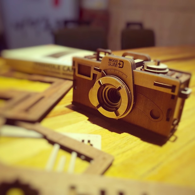 A wooden model of a vintage camera is placed on a table with blurred objects surrounding it. The focus is on the intricately designed front of the camera, displaying precision-cut details and engraved markings. The warm lighting casts soft shadows, highlighting the texture of the wood.
