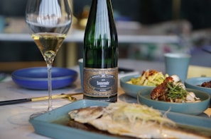 A wine bottle and a filled glass are positioned on a dining table. The table features a selection of dishes, including a plate with a grilled fish in the foreground and side dishes in blue bowls. The setting suggests an elegant meal.