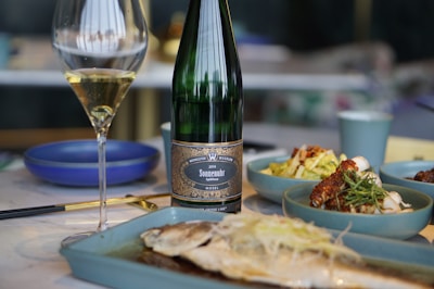 A wine bottle and a filled glass are positioned on a dining table. The table features a selection of dishes, including a plate with a grilled fish in the foreground and side dishes in blue bowls. The setting suggests an elegant meal.