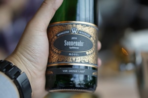 A hand holding a green glass bottle of wine with a detailed label. The label is ornate with gold patterns and text indicating the wine type, vintage year, and origin from Mosel. The wrist of the person is adorned with a black digital watch. The background is softly blurred, suggesting a casual or relaxed setting.