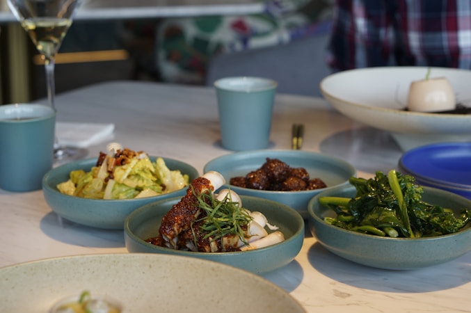 A selection of small dishes featuring a variety of foods, including leafy greens, a noodle-like dish topped with red sauce and garnished with herbs, a dish with cooked greens, and a bowl of mixed salad with lettuce. The dishes are placed on top of a white marble table. In the background, there is a wine glass, a cup, and a partially visible person wearing a plaid shirt.