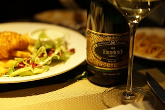 A dinner table setting features a plate with a serving of fish and chips accompanied by a side salad with leafy greens and dressing. Next to the plate is a wine bottle labeled 'Himmelreich Spätlese Mosel' from the year 2012. A wine glass filled with white wine is placed in front of the bottle, creating a cozy and elegant dining atmosphere.