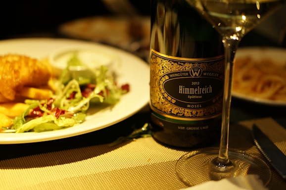 A dinner table setting features a plate with a serving of fish and chips accompanied by a side salad with leafy greens and dressing. Next to the plate is a wine bottle labeled 'Himmelreich Spätlese Mosel' from the year 2012. A wine glass filled with white wine is placed in front of the bottle, creating a cozy and elegant dining atmosphere.