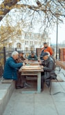 A group of elderly men gather around a wooden table outdoors, engaged in a board game. The setting is a park area with benches and trees, and a historical building is visible in the background. The scene is peaceful, with autumn leaves scattered around, and the men appear focused and relaxed.