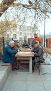 A group of elderly men gather around a wooden table outdoors, engaged in a board game. The setting is a park area with benches and trees, and a historical building is visible in the background. The scene is peaceful, with autumn leaves scattered around, and the men appear focused and relaxed.