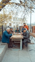 A group of elderly men gather around a wooden table outdoors, engaged in a board game. The setting is a park area with benches and trees, and a historical building is visible in the background. The scene is peaceful, with autumn leaves scattered around, and the men appear focused and relaxed.