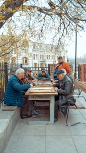 A group of elderly men gather around a wooden table outdoors, engaged in a board game. The setting is a park area with benches and trees, and a historical building is visible in the background. The scene is peaceful, with autumn leaves scattered around, and the men appear focused and relaxed.
