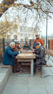 A group of elderly men gather around a wooden table outdoors, engaged in a board game. The setting is a park area with benches and trees, and a historical building is visible in the background. The scene is peaceful, with autumn leaves scattered around, and the men appear focused and relaxed.