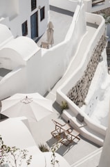 A serene rooftop terrace features whitewashed architectural elements, including curved walls and staircases. A large white umbrella shades two wooden director-style chairs, and there is a small table nearby. The space is bordered by a stone wall, and there are touches of greenery scattered around.