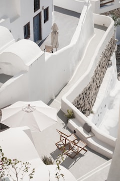 A serene rooftop terrace features whitewashed architectural elements, including curved walls and staircases. A large white umbrella shades two wooden director-style chairs, and there is a small table nearby. The space is bordered by a stone wall, and there are touches of greenery scattered around.