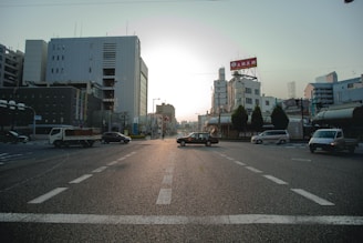 An urban street scene with multiple vehicles at an intersection. Surrounding the road are modern buildings with signage, and trees line part of the street. The lighting suggests either sunrise or sunset, creating an ambient glow.