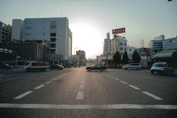 An urban street scene with multiple vehicles at an intersection. Surrounding the road are modern buildings with signage, and trees line part of the street. The lighting suggests either sunrise or sunset, creating an ambient glow.